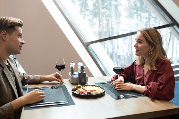 Giovane uomo elegante in smart casuawlear guardando la sua ragazza mentre tiene in mano un bicchiere di vino rosso e parla con lei a cena al ristorante