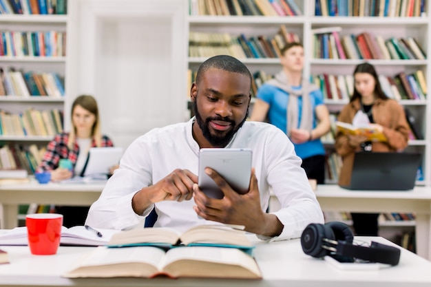 Giovane uomo felice africano bello, studente, con la barba e in camicia bianca, seduto al tavolo con i libri nella sala di lettura delle biblioteche