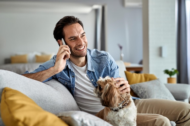 Giovane uomo felice che si diverte con il suo cane a casa e comunica tramite telefono cellulare