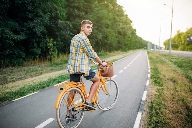 Giovane uomo felice sulla bicicletta vintage gialla con cestino, parco estivo verde. Escursioni in bicicletta all'aperto. Persona di sesso maschile sulla bici retrò