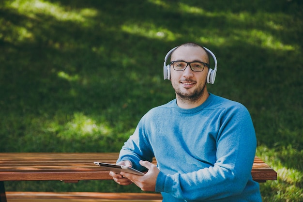 Giovane uomo felice, uomo d'affari o studente in occhiali casual camicia blu seduto al tavolo con le cuffie, tablet pc nel parco cittadino, ascoltare musica, riposare all'aperto sulla natura verde. Concetto di tempo libero di stile di vita.