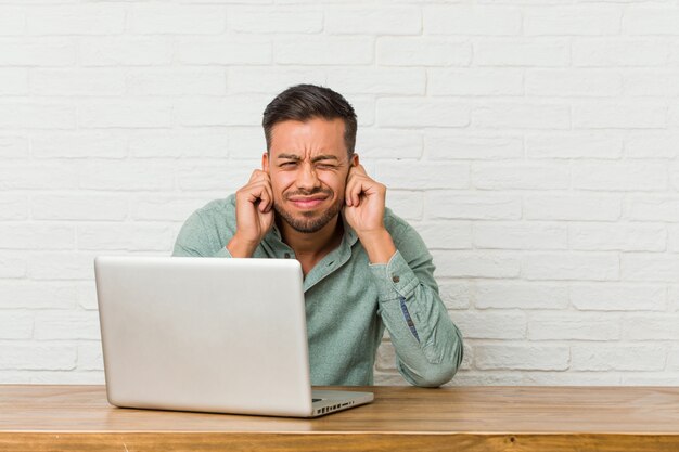 Giovane uomo filippino seduto a lavorare con il suo laptop che copre le orecchie con le mani.