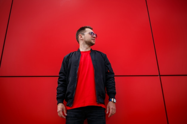 Giovane uomo gioioso con la barba con un sorriso positivo in una t-shirt in una giacca nera alla moda in una t-shirt in posa vicino a un edificio di metallo rosso in città. Ragazzo urbano felice sulla strada. Abbigliamento da uomo