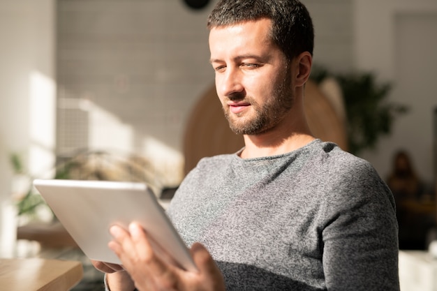 Giovane uomo in abbigliamento casual guardando qualcosa online mentre trascorre il tempo nella caffetteria in giornata di sole