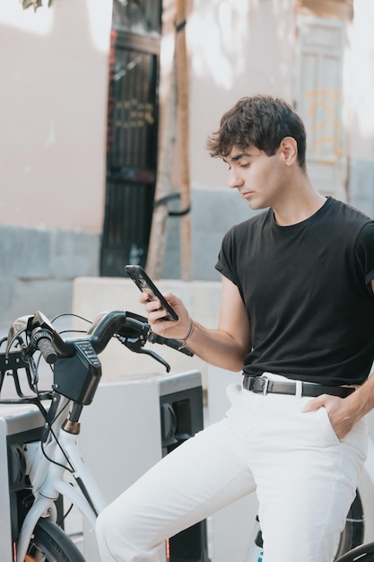 Giovane uomo in abiti moderni che usa il suo telefono vicino a un servizio di noleggio biciclette, sblocca biciclette a noleggio per spostarsi in città, cerca telefono, servizi di noleggio, scooter, bici e auto, stile di vita in città