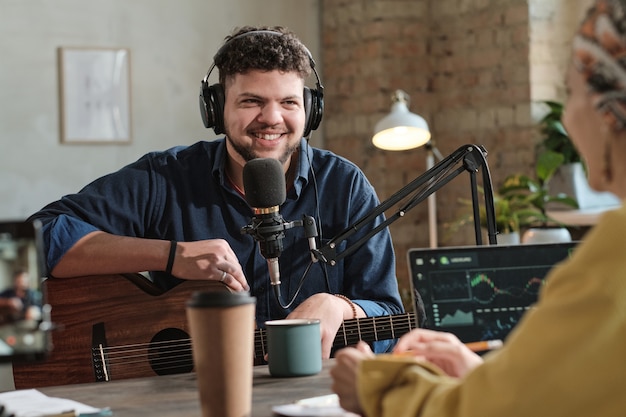 Giovane uomo in cuffia seduto con la chitarra e dando un'intervista a radio dj in studio