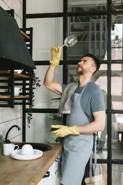 giovane uomo in grembiule e guanti fa le faccende domestiche lava i piatti con un asciugamano sulla spalla uomini lavori domestici aiuto domestico in una cucina elegante in un appartamento moderno