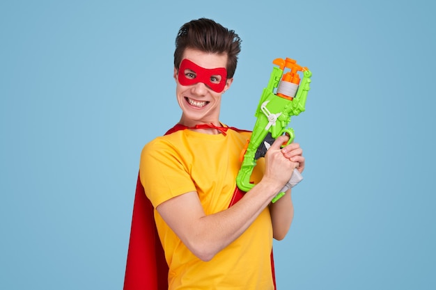 Giovane uomo in maschera da supereroe e mantello sorridente per la fotocamera e tirando il grilletto su un'arma giocattolo su sfondo blu