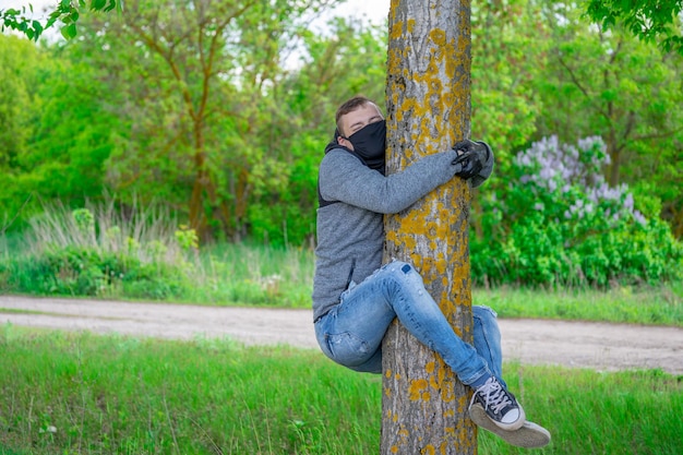 Giovane uomo in maschera medica nera e guanti appesi all'albero in un bosco Maschio adulto che si gode la natura in campagna nel periodo dell'infezione epidemica di coronavirus