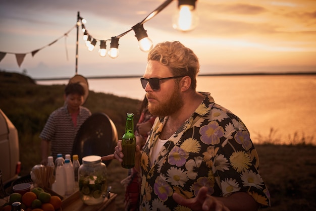 Giovane uomo in occhiali da sole che tiene in mano una bottiglia di birra e balla con i suoi amici alla festa in spiaggia all'aperto