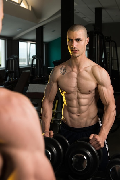 Giovane uomo in piedi forte davanti a uno specchio e flettendo i muscoli Modello di fitness bodybuilder atletico muscolare in posa dopo gli esercizi