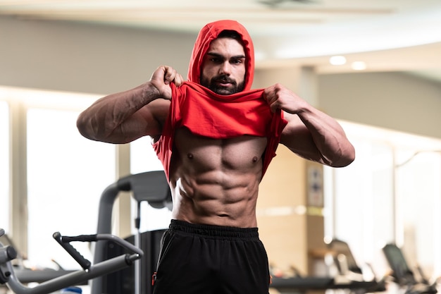 Giovane uomo in piedi forte in palestra e flettendo i muscoli in una felpa con cappuccio rossa muscoloso bodybuilder atletico modello di fitness in posa dopo gli esercizi