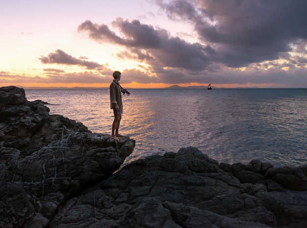 Giovane uomo in piedi sulle rocce e rivolto verso l'oceano al tramonto TimeOutdoor ConceptCopy Space