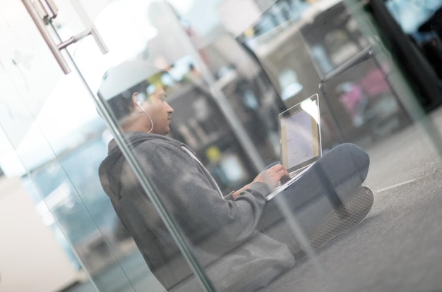 Giovane uomo indiano sviluppatore di software che utilizza un computer portatile per scrivere codice di programmazione mentre è seduto sul pavimento in un moderno ufficio di avvio creativo