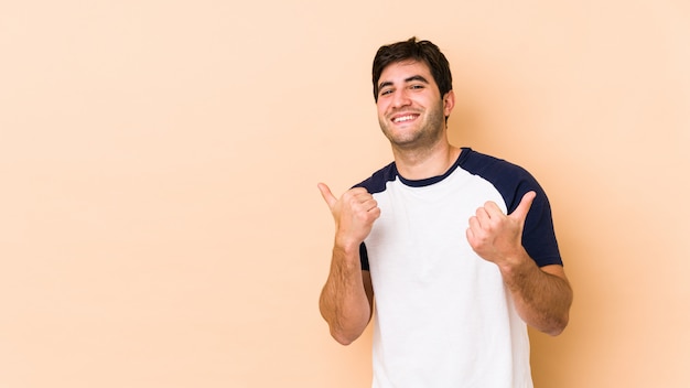 Giovane uomo isolato su beige alzando entrambi i pollici, sorridendo e confidont.