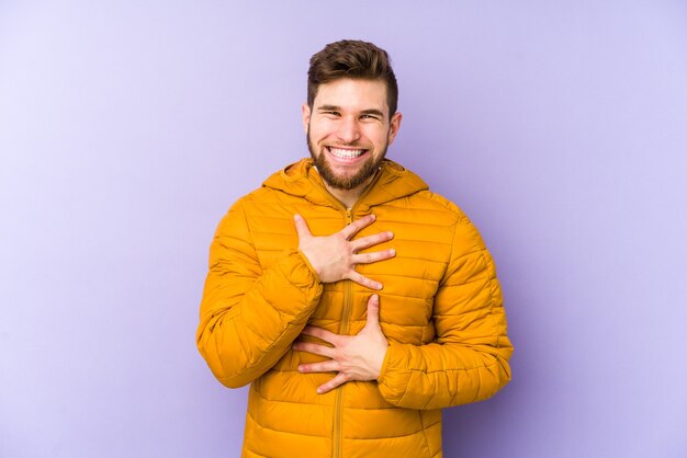 Giovane uomo isolato su sfondo viola ride allegramente e si diverte a tenere le mani sullo stomaco.