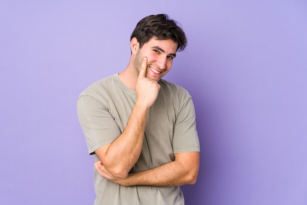 Giovane uomo isolato su sfondo viola sorridendo felice e fiducioso, toccando il mento con la mano.