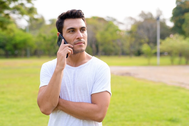 Giovane uomo ispanico bello pensare e parlare al telefono al parco