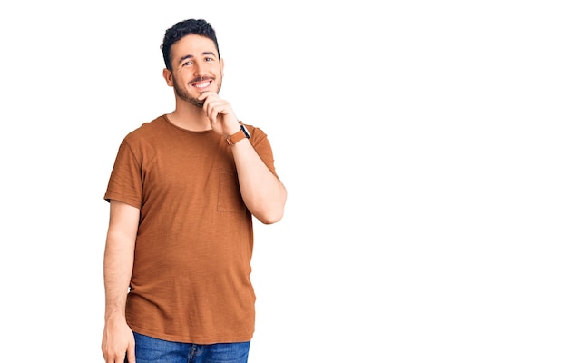 Giovane uomo ispanico che indossa abiti casual che guarda fiducioso alla macchina fotografica con il sorriso con le braccia incrociate e la mano alzata sul mento pensando positivo