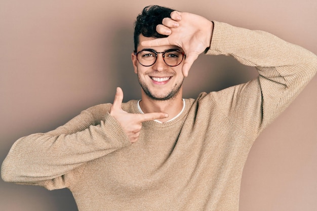 Giovane uomo ispanico che indossa abiti casual e occhiali sorridenti facendo cornice con le mani e le dita con la creatività del viso felice e il concetto di fotografia