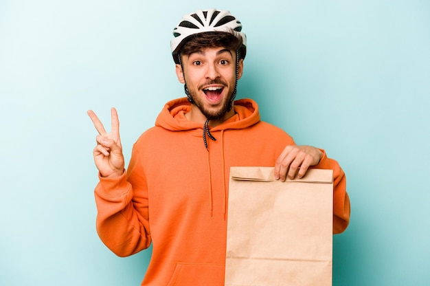 Giovane uomo ispanico che indossa un casco bici con in mano un cibo da asporto isolato su sfondo blu gioioso e spensierato che mostra un simbolo di pace con le dita