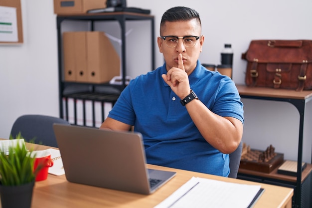 Giovane uomo ispanico che lavora in ufficio con il computer portatile che chiede di stare zitto con il dito sulle labbra. silenzio e concetto segreto.