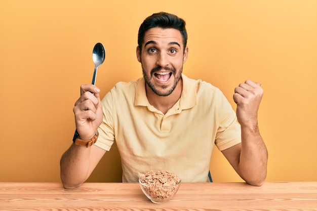 Giovane uomo ispanico che mangia cereali integrali sani celears seduto sul tavolo che punta il pollice verso l'alto sorridendo felice con la bocca aperta