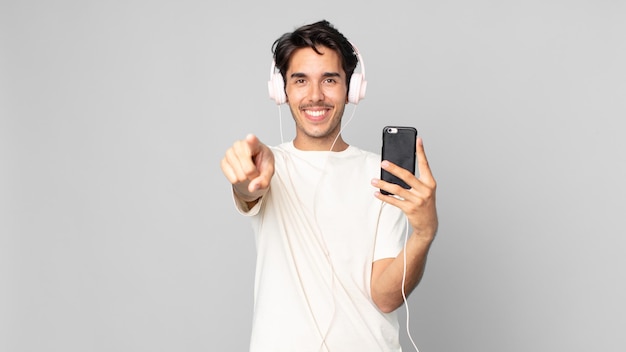 Giovane uomo ispanico che punta alla telecamera scegliendo te con cuffie e smartphone