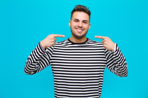 Giovane uomo ispanico che sorride con confidenza indicando il proprio ampio sorriso, atteggiamento positivo, rilassato, soddisfatto contro la parete blu