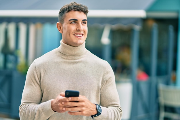 Giovane uomo ispanico che sorride felice utilizzando lo smartphone in città.