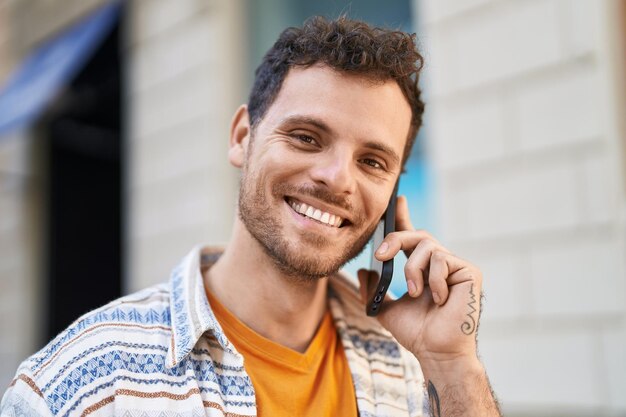 Giovane uomo ispanico che sorride fiducioso parlando sullo smartphone in strada