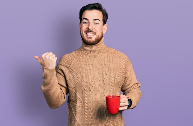 Giovane uomo ispanico che tiene il caffè che punta il pollice verso l'alto sorridendo felice con la bocca aperta