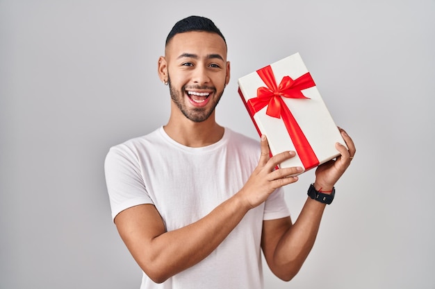 Giovane uomo ispanico che tiene regali che festeggiano pazzi e stupiti per il successo con gli occhi aperti che urlano eccitati.