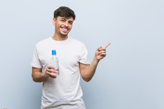 Giovane uomo ispanico che tiene una bottiglia di acqua che sorride allegramente indicando con l'indice di distanza.