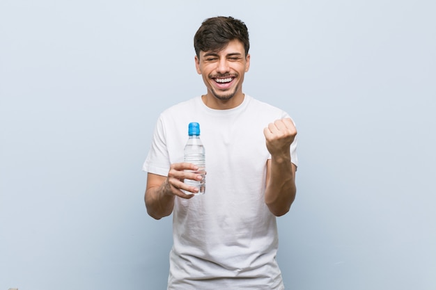 Giovane uomo ispanico che tiene una bottiglia di acqua incoraggiante spensierata ed eccitata. Concetto di vittoria.