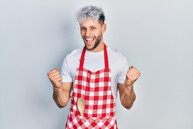 Giovane uomo ispanico con capelli tinti moderni che indossa un grembiule che celebra sorpreso e stupito per il successo con le braccia alzate e il concetto di vincitore degli occhi aperti