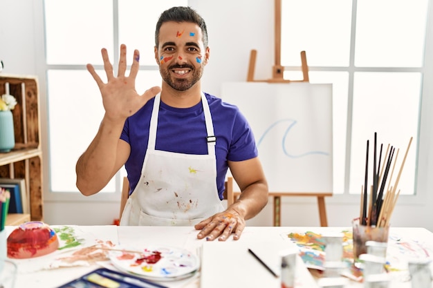 Giovane uomo ispanico con la barba allo studio d'arte con la faccia dipinta che mostra e punta verso l'alto con le dita numero cinque mentre sorride fiducioso e felice