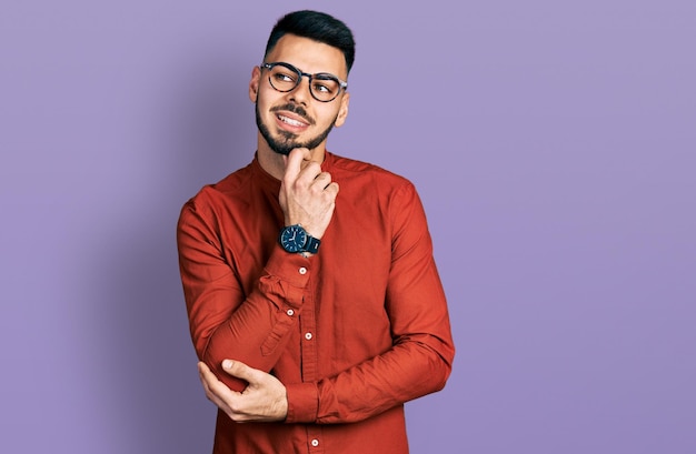 Giovane uomo ispanico con la barba che indossa camicia da lavoro e occhiali con la mano sul mento pensando alla domanda espressione pensierosa sorridente con il concetto di dubbio viso premuroso