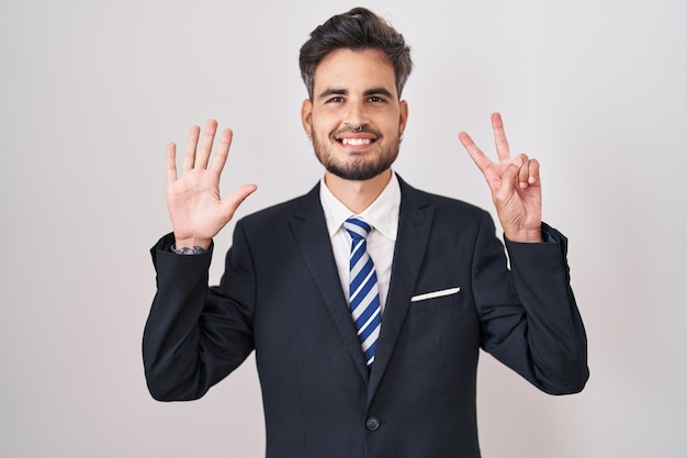 Giovane uomo ispanico con tatuaggi che indossa tailleur e cravatta mostrando e puntando verso l'alto con le dita numero sette mentre sorride fiducioso e felice