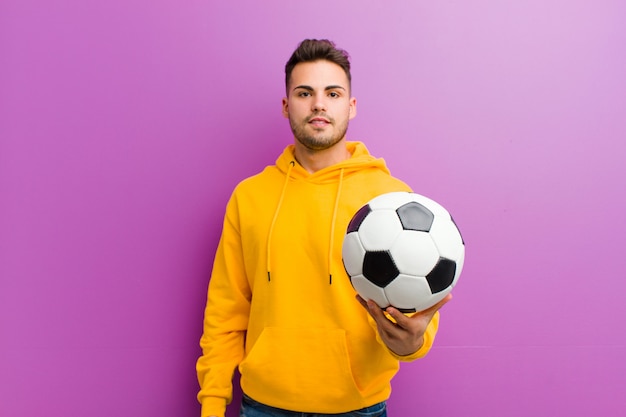 Giovane uomo ispanico con un pallone da calcio contro la parete viola