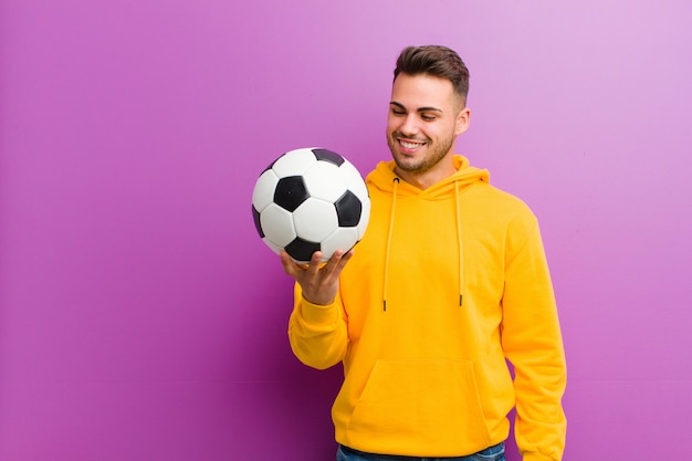 Giovane uomo ispanico con un pallone da calcio su sfondo viola