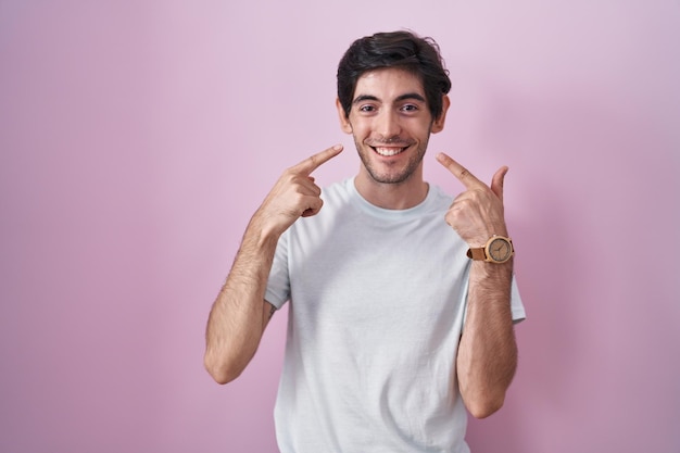 Giovane uomo ispanico in piedi su sfondo rosa sorridente mostrando allegro e indicando con le dita i denti e la bocca. concetto di salute dentale.