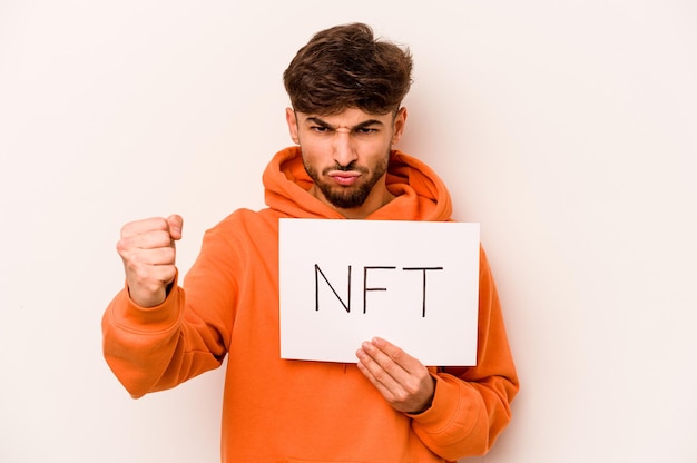 Giovane uomo ispanico in possesso di un cartello NFT isolato su sfondo bianco che mostra il pugno alla fotocamera espressione facciale aggressiva