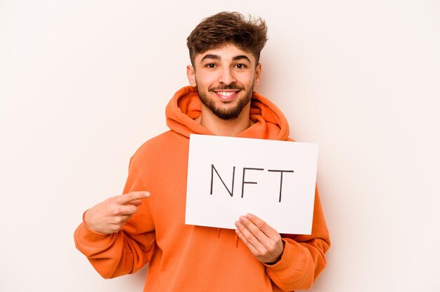 Giovane uomo ispanico in possesso di un cartello NFT isolato su sfondo bianco sorridente e indicando da parte mostrando qualcosa in uno spazio vuoto