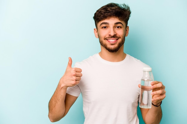 Giovane uomo ispanico in possesso di una bottiglia d'acqua isolata su sfondo bianco sorridente e alzando il pollice