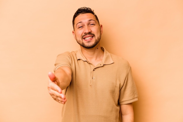 Giovane uomo ispanico isolato su sfondo beige che allunga la mano alla telecamera nel gesto di saluto