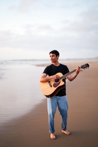 giovane uomo latino che suona la chitarra a piedi nudi felice sulla riva della spiaggia al tramonto