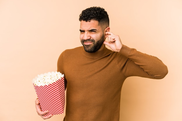 Giovane uomo latino che tiene un pop corn isolato che copre le orecchie con le mani.