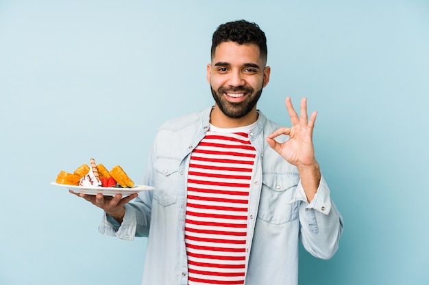 Giovane uomo latino che tiene un waffle allegro e fiducioso mostrando gesto ok.