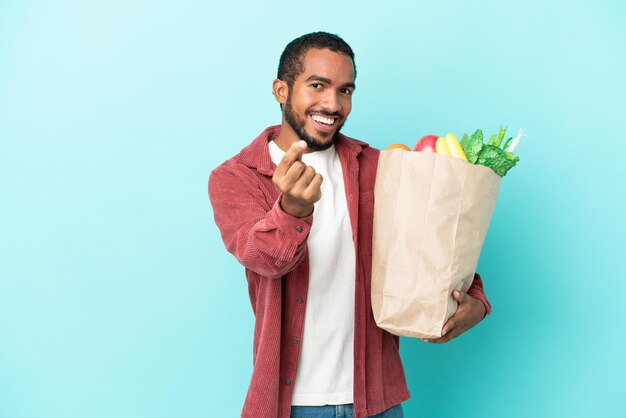 Giovane uomo latino che tiene una borsa della spesa isolata su fondo blu che fa il gesto dei soldi